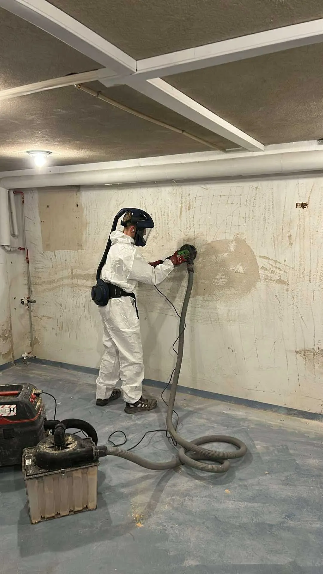 Medarbejder i heldragt og friskluftmaske sliber betonvaeg i kaelder med stoevsug